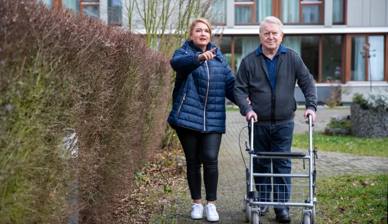 Frau hilft Mann, der mit Rollator neben ihr läuft