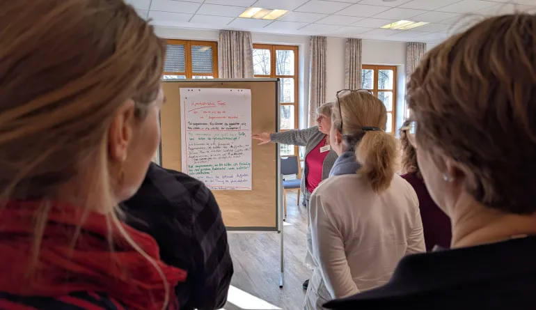 eine Referentin erklärt was an der Stellwand, der Blick geht an zwei Teilnehmenden vorbei