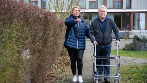 Frau hilft Mann, der mit Rollator neben ihr läuft